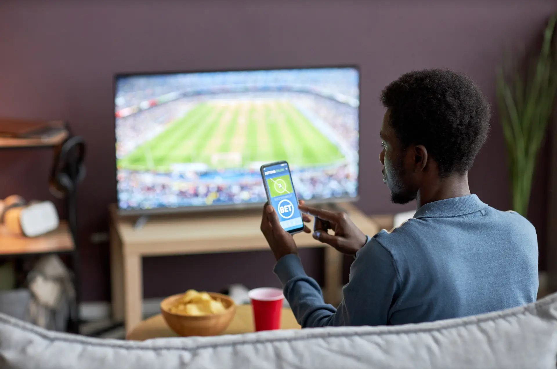 Person placing a football bet on a smartphone while watching a live soccer match.