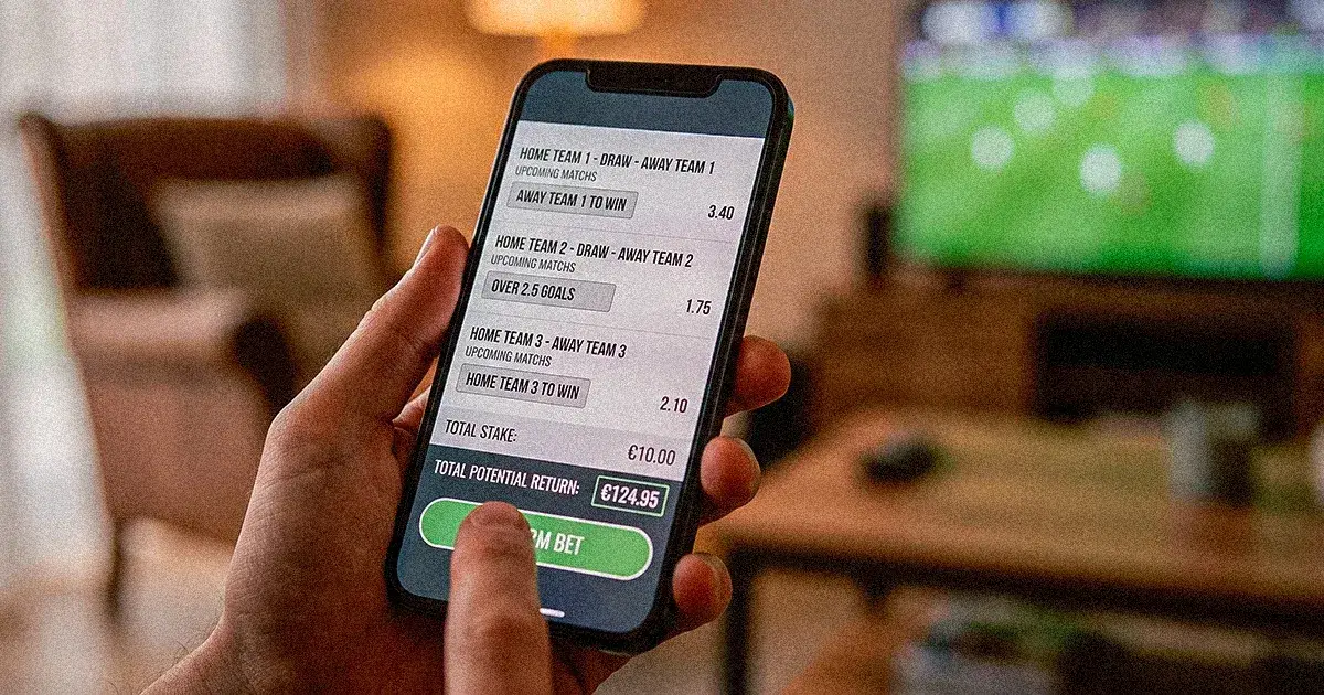 Mano sostiene un celular con un boleto de apuestas deportivas frente a la televisión
